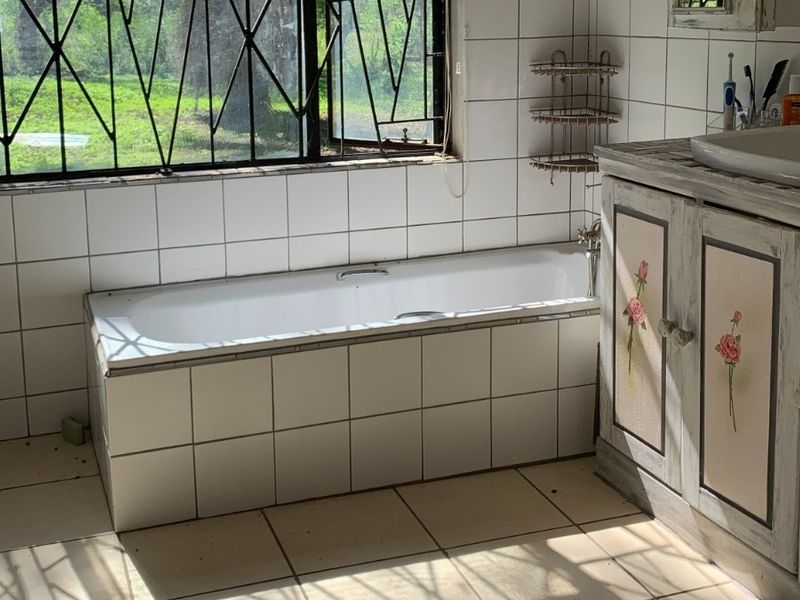 A dated bathroom featuring a built-in tiled bathtub, a vanity cabinet with basin, and large windows with security bars overlooking the garden.