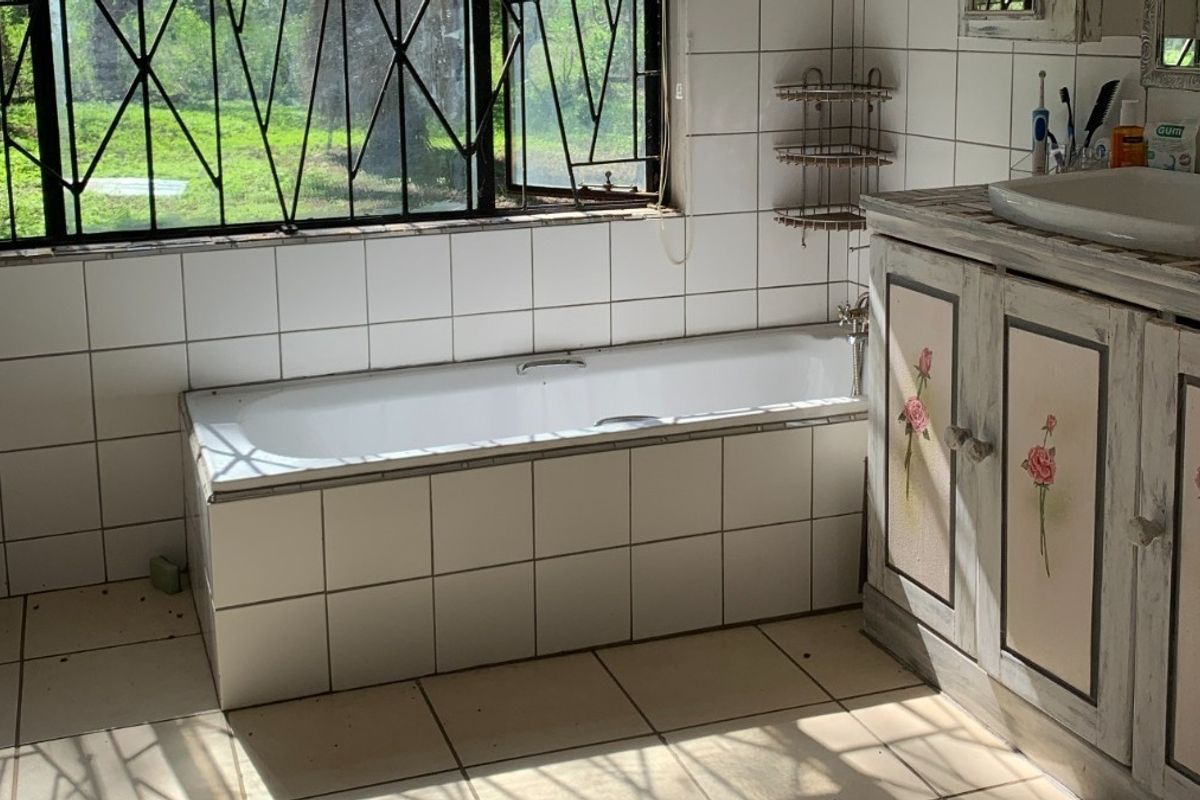 A dated bathroom featuring a built-in tiled bathtub, a vanity cabinet with basin, and large windows with security bars overlooking the garden.