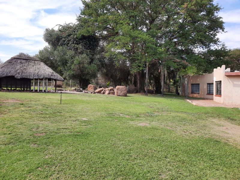 Spacious garden area featuring a traditional thatched lapa, mature trees, and natural stone landscaping adjacent to the main dwelling.