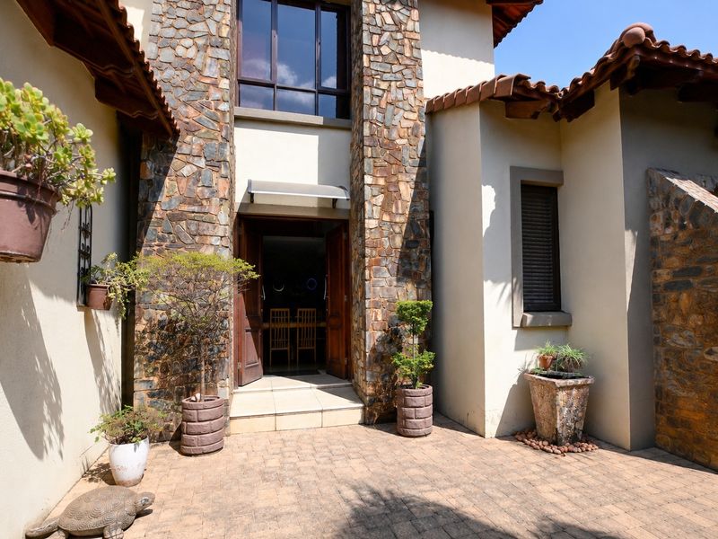 A welcoming entrance featuring stone-clad pillars, terracotta roof tiles, and a paved courtyard leading to double wooden doors.
