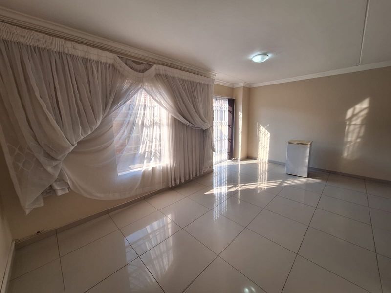 A bright, open-plan living area featuring neutral tiled flooring, large windows with curtains, and a security gate leading to the exterior.