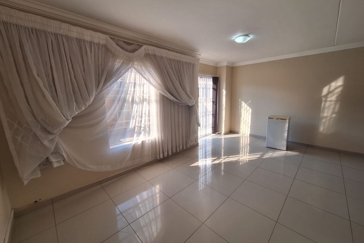 A bright, open-plan living area featuring neutral tiled flooring, large windows with curtains, and a security gate leading to the exterior.