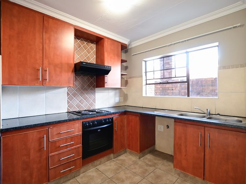 A functional kitchen featuring wooden cabinetry, dark countertops, built-in oven and hob, tiled backsplash, and a double bowl sink with natural light.