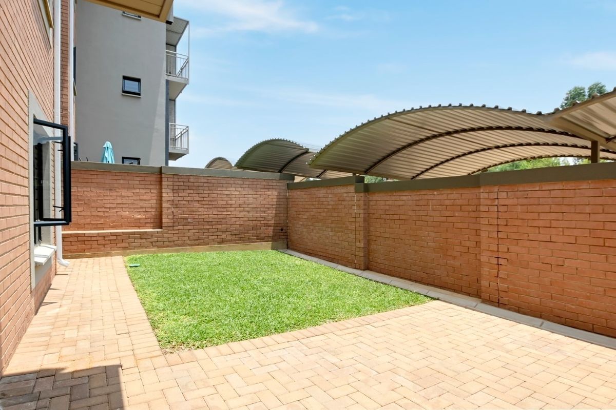 A private rear courtyard featuring a paved patio area, a small lawn, and brick boundary walls with an overhead carport structure.