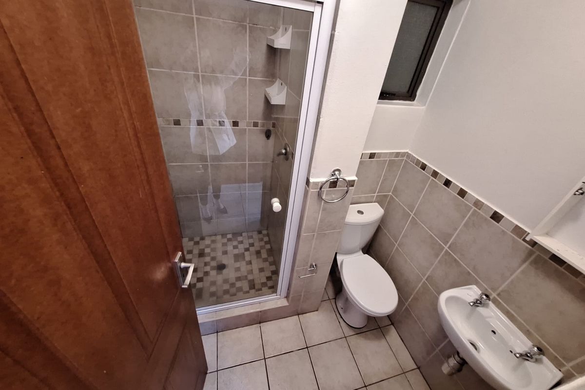 A functional bathroom featuring a tiled shower enclosure, pedestal basin, and toilet with neutral wall tiling and a small window for ventilation.