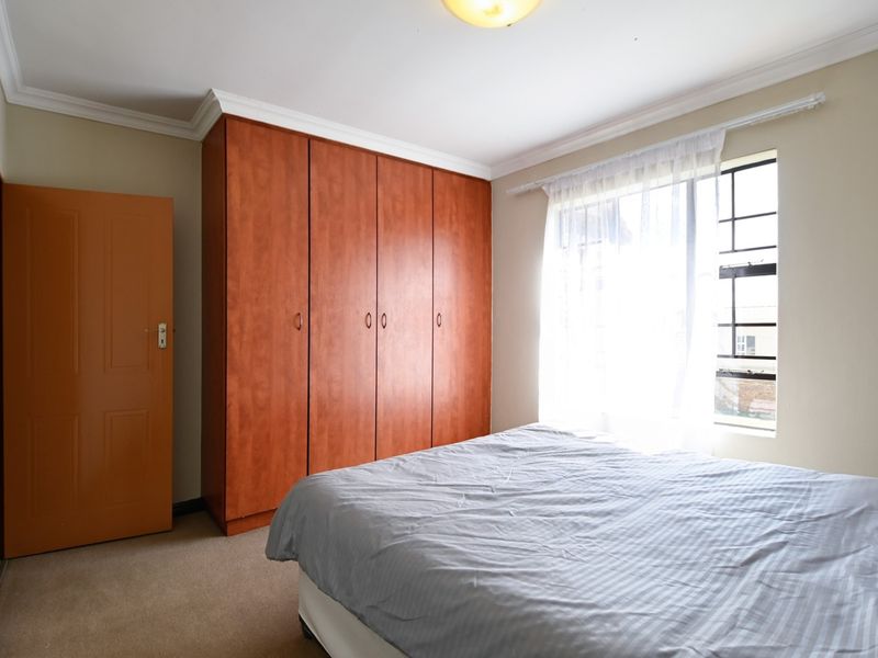 A bedroom featuring built-in wooden cupboards, neutral wall finishes, and a window with security bars, providing a functional and well-lit living space.