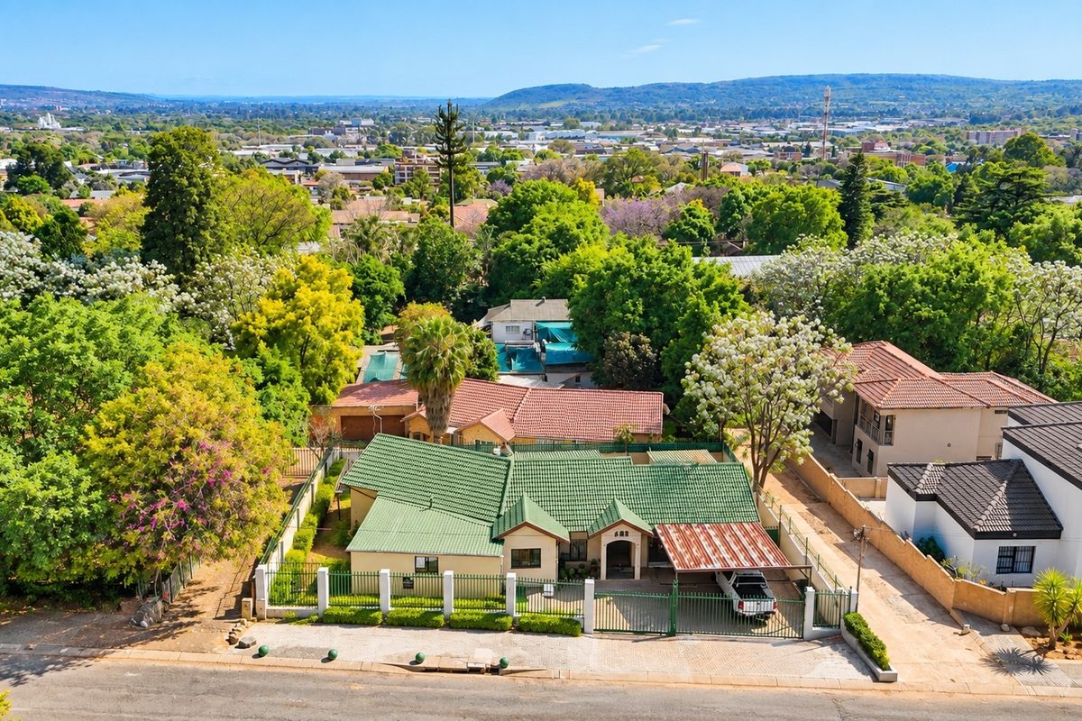 Aerial view of a residential property featuring a green tiled roof, perimeter fencing, and a carport, set within a lush, established suburban neighborhood.