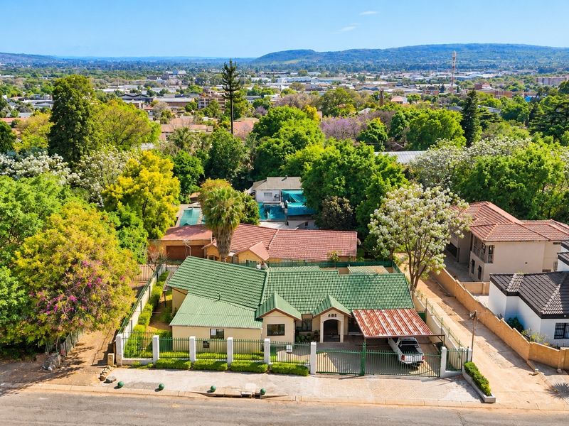 Aerial view of a residential property featuring a green tiled roof, perimeter fencing, and a carport, set within a lush, established suburban neighborhood.