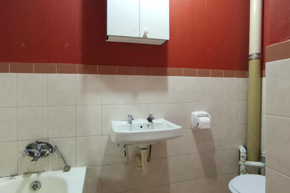 A functional bathroom featuring a wall-mounted basin, bathtub, and toilet, with dated finishes and exposed plumbing requiring cosmetic updates.