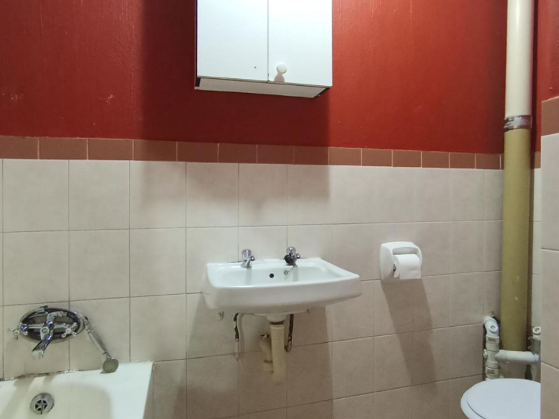 A functional bathroom featuring a wall-mounted basin, bathtub, and toilet, with dated finishes and exposed plumbing requiring cosmetic updates.