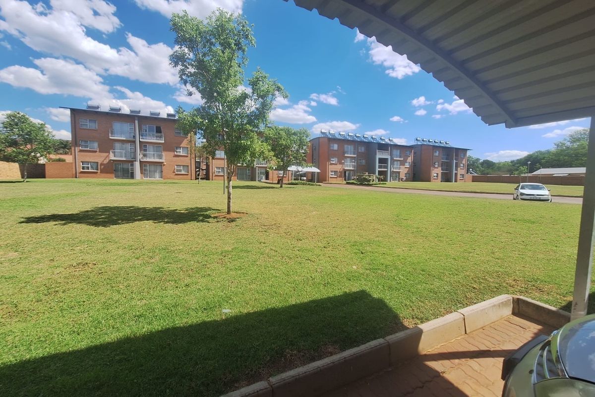 View of the communal garden and brick apartment complex from a covered parking area, featuring well-maintained lawns and solar water heating installations.