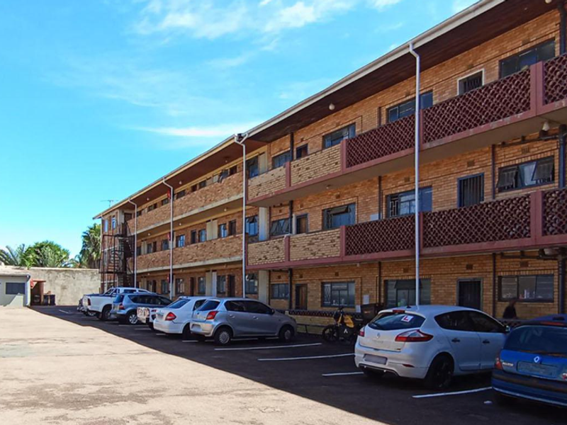 Exterior view of a multi-story apartment building featuring brick facade, private balconies with decorative screens, and an adjacent asphalt parking area.