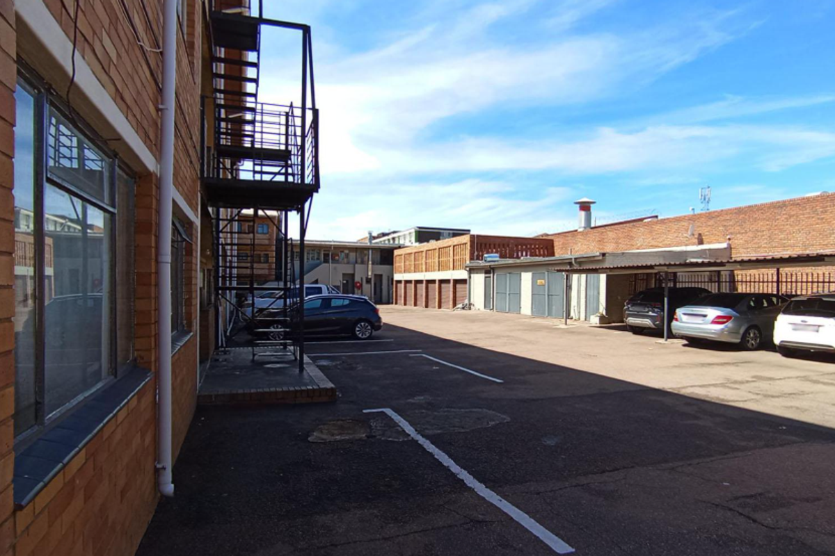 Rear exterior view of the apartment building featuring a brick facade, metal fire escape, and an asphalt parking area with covered carports.