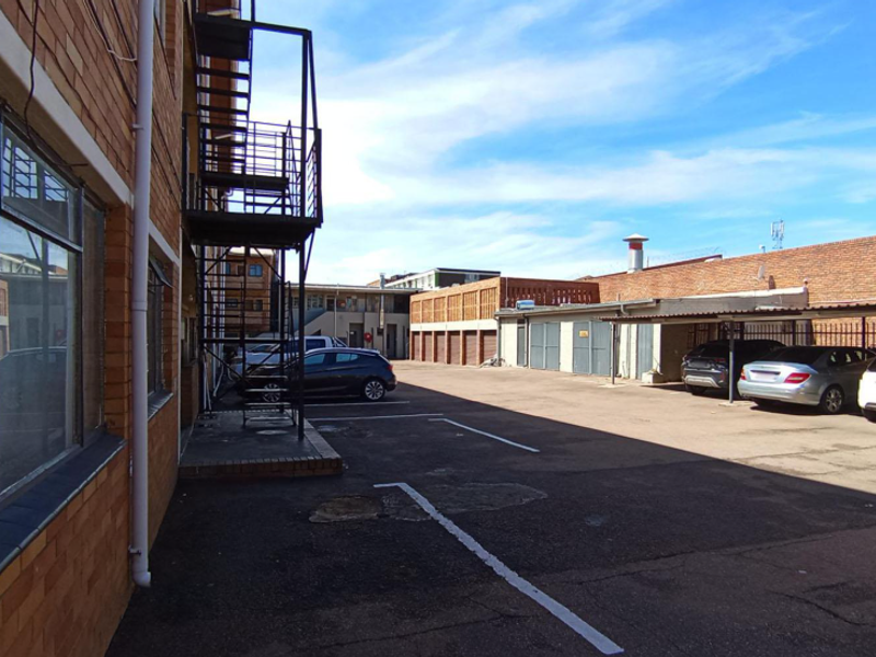 Rear exterior view of the apartment building featuring a brick facade, metal fire escape, and an asphalt parking area with covered carports.