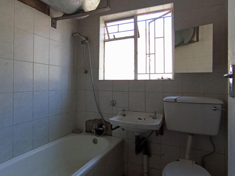 A dated bathroom featuring a bathtub, wall-mounted basin, and toilet, requiring renovation to address damaged tiling and exposed plumbing fixtures.