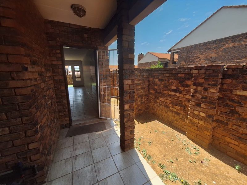 A covered patio area featuring exposed brick walls, tiled flooring, and a security gate, leading into the interior of the townhouse.