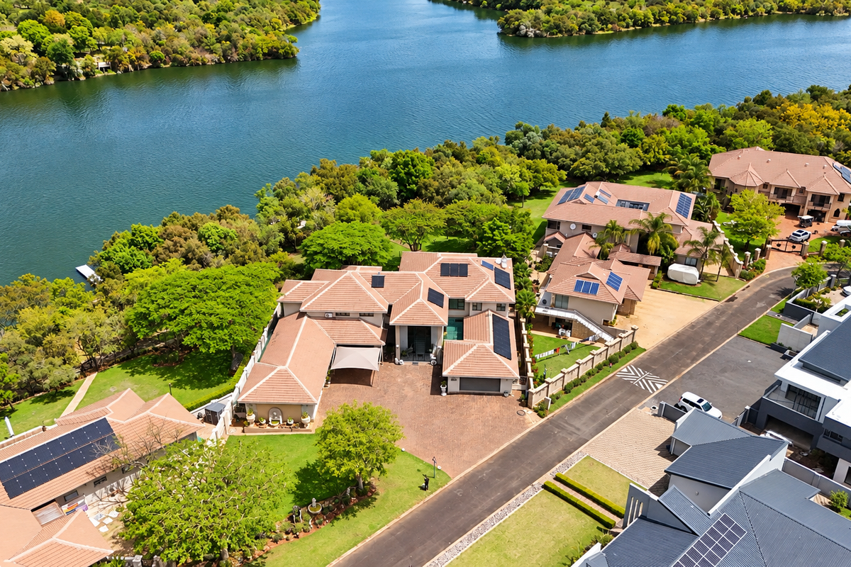 Aerial view of a large residential property featuring multiple tiled roof structures, solar panels, a paved driveway, and extensive landscaped gardens along the waterfront.