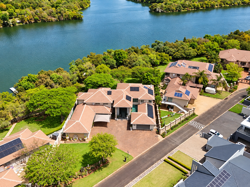 Aerial view of a large residential property featuring multiple tiled roof structures, solar panels, a paved driveway, and extensive landscaped gardens along the waterfront.