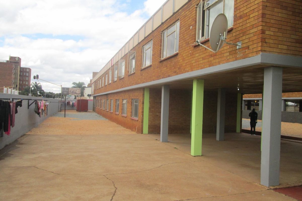 Rear exterior view of a multi-story brick apartment building featuring a covered ground-level area with concrete pillars and paved outdoor space.