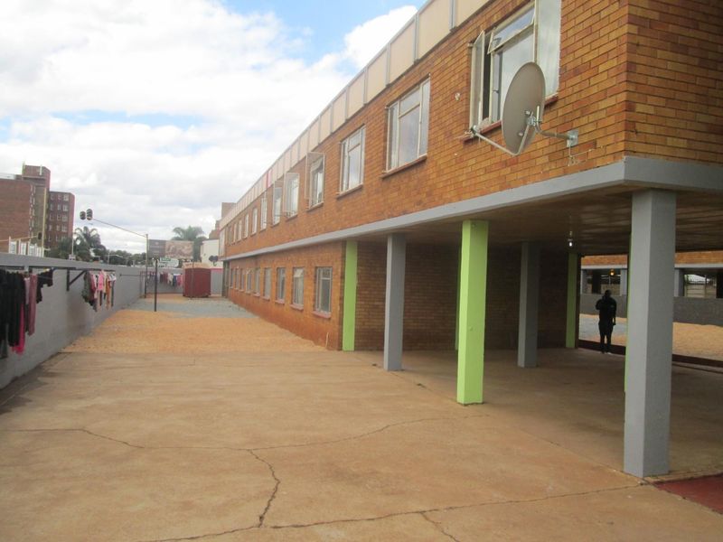 Rear exterior view of a multi-story brick apartment building featuring a covered ground-level area with concrete pillars and paved outdoor space.