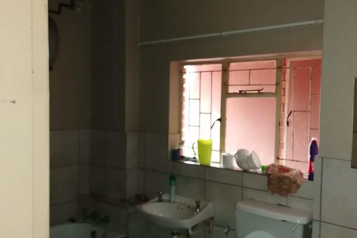A dated bathroom featuring a standard bathtub, pedestal basin, and toilet, with security bars on the window and tiled splashback areas.