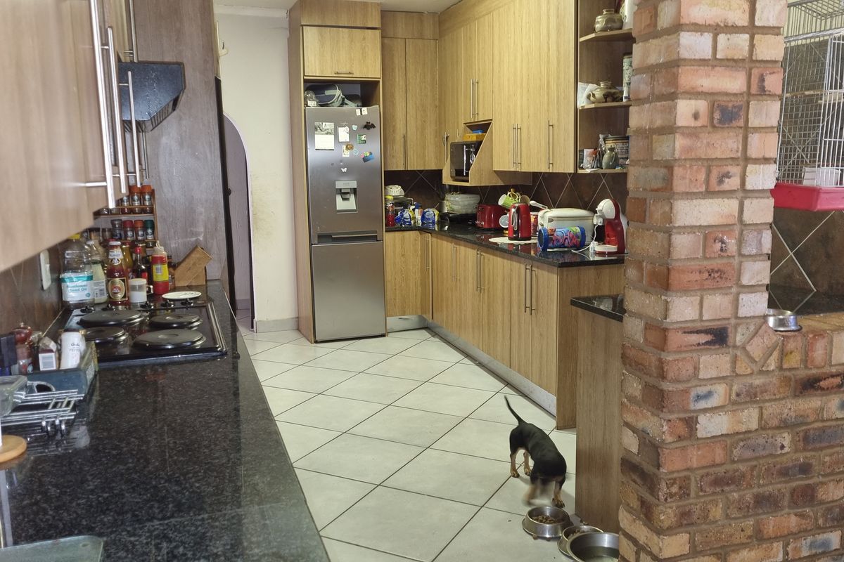 A functional kitchen featuring wood-look cabinetry, dark granite countertops, a built-in hob, and tiled flooring, with an open-plan layout and exposed brick pillar.