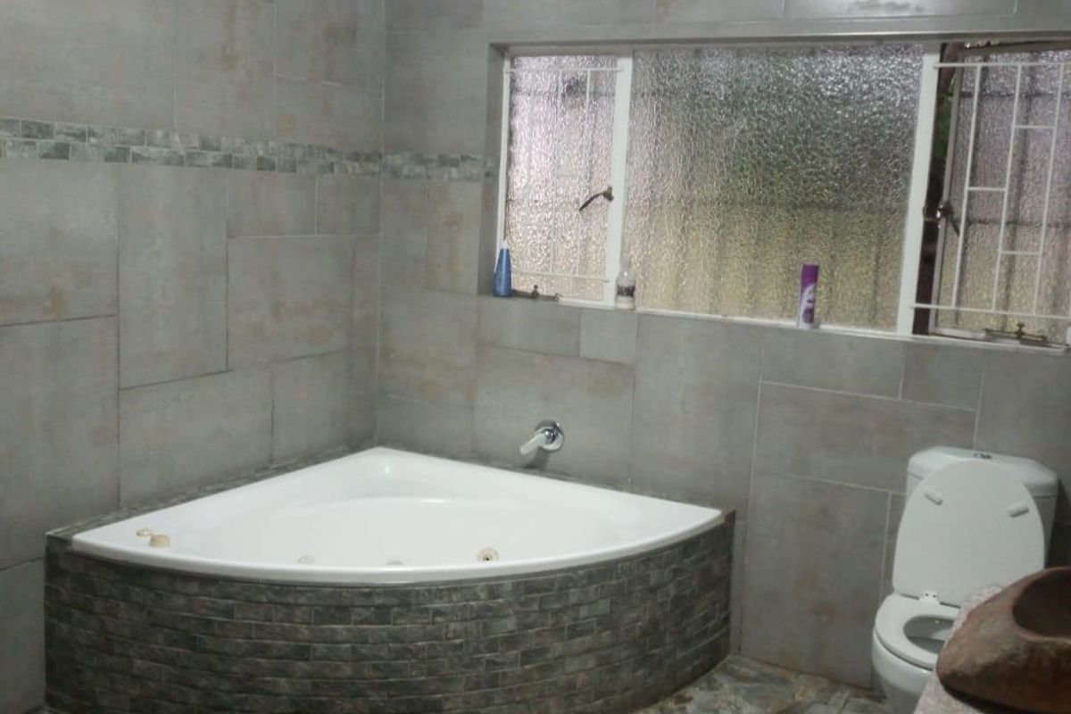 A bathroom featuring a corner bathtub with stone-look tiling, a toilet, and tiled wall finishes with security bars on the window.
