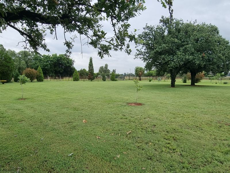 A spacious, well-maintained garden area featuring expansive green lawns, mature trees, and perimeter fencing on this small holding property.