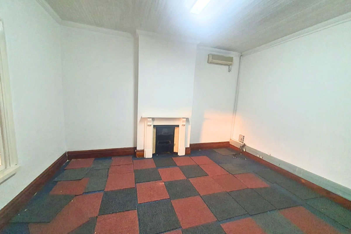 An empty living room featuring a fireplace, mantelpiece, and wall-mounted air conditioning unit, finished with dated, worn carpet tiles.