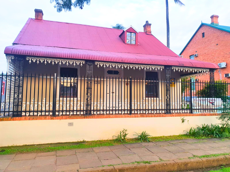 A charming historic-style building featuring a red corrugated metal roof, decorative wrought iron veranda detailing, and a secure front boundary fence.
