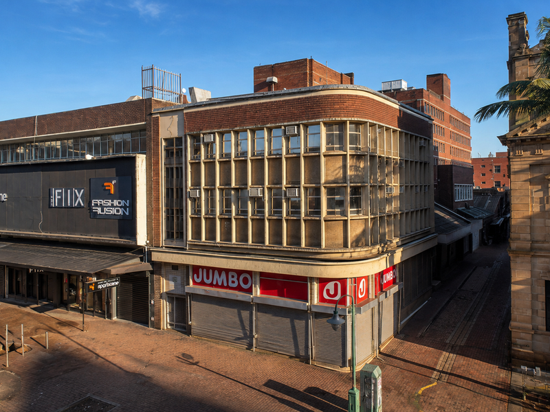 Street-level view of a multi-story commercial building featuring brick facade, metal roller shutters, and integrated retail signage in an urban setting.