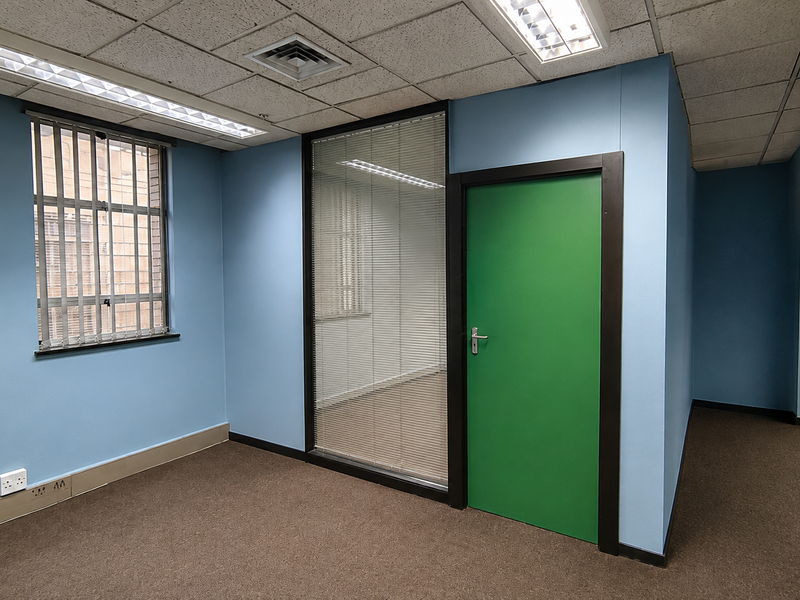 A vacant commercial office space featuring blue walls, carpeted flooring, a glass partition, and a green internal door with integrated lighting and air conditioning.