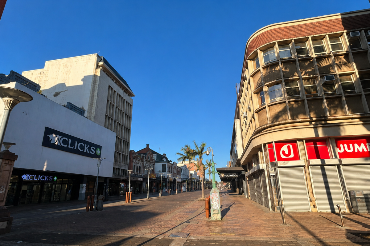 A street-level view of a commercial pedestrian area featuring retail storefronts with security shutters and multi-story building facades under a clear sky.