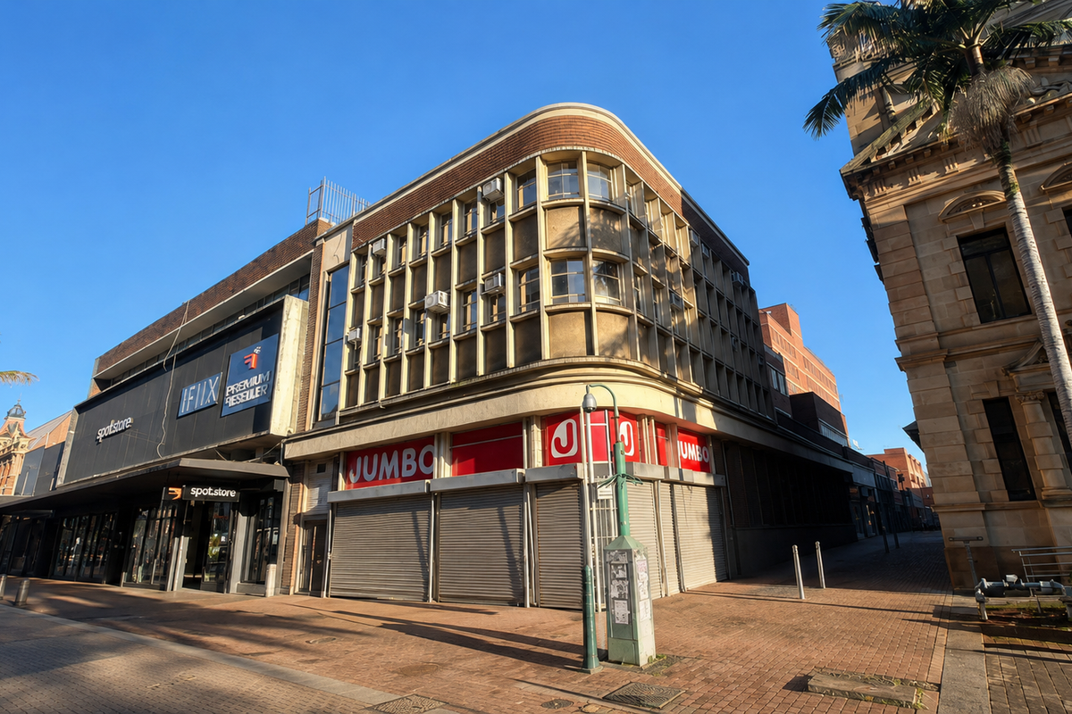 Street-level view of a commercial building facade featuring retail storefronts with roller shutters and prominent signage in a pedestrian-friendly urban setting.