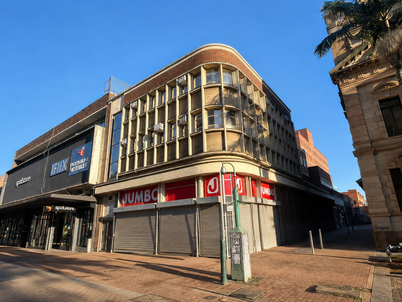 Street-level view of a commercial building facade featuring retail storefronts with roller shutters and prominent signage in a pedestrian-friendly urban setting.