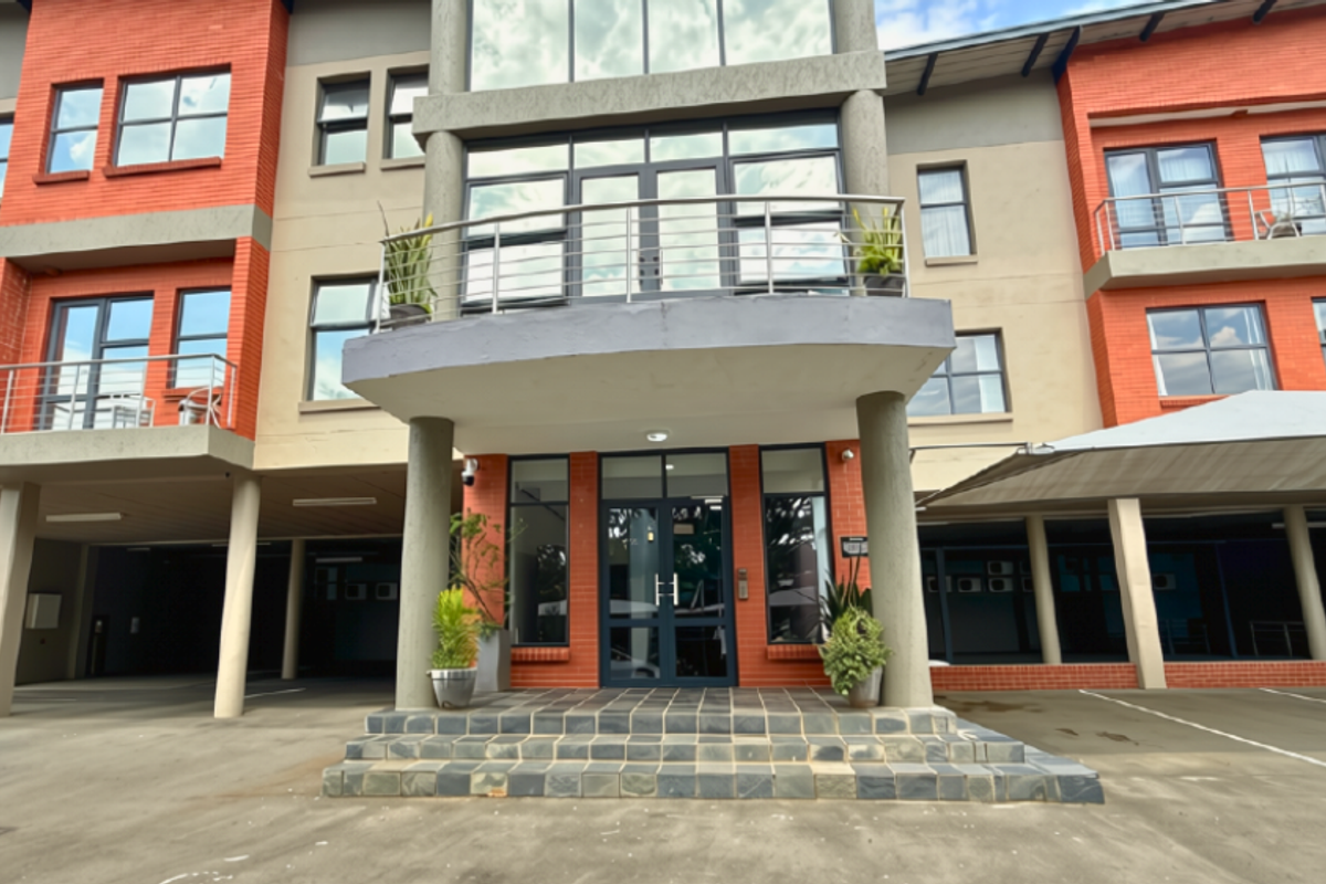Front elevation of a modern commercial building featuring brick accents, glass entrance doors, balconies with stainless steel railings, and a covered parking area.