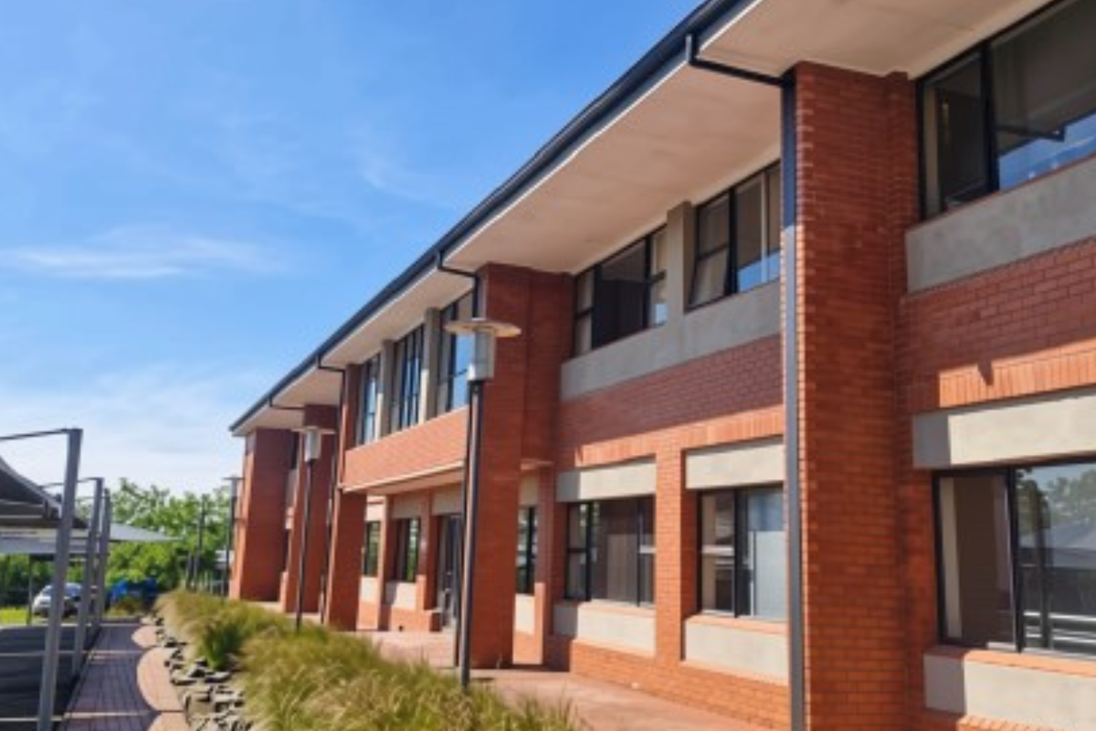 A modern two-story office building featuring a red brick facade, large aluminium-framed windows, and an adjacent covered parking area.