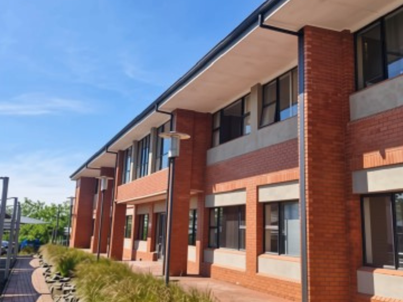 A modern two-story office building featuring a red brick facade, large aluminium-framed windows, and an adjacent covered parking area.