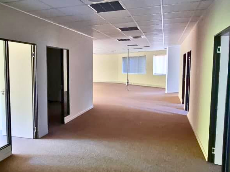 Spacious, empty open-plan office area featuring a suspended ceiling, recessed lighting, and carpeted flooring with multiple internal office access points.