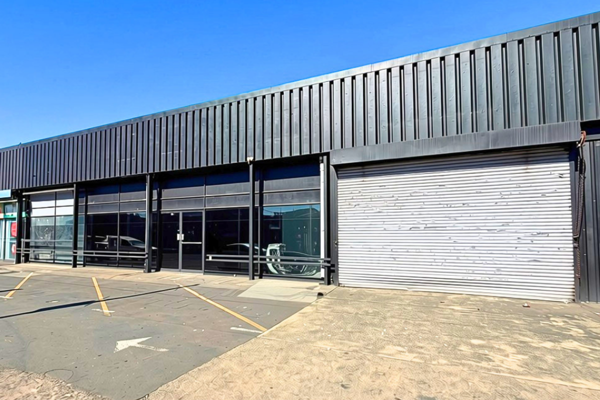 Industrial storefront featuring a large metal roller shutter door, glass facade, and corrugated metal cladding with an adjacent asphalt parking area.
