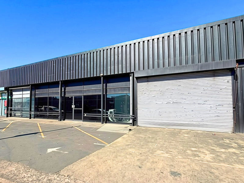 Industrial storefront featuring a large metal roller shutter door, glass facade, and corrugated metal cladding with an adjacent asphalt parking area.