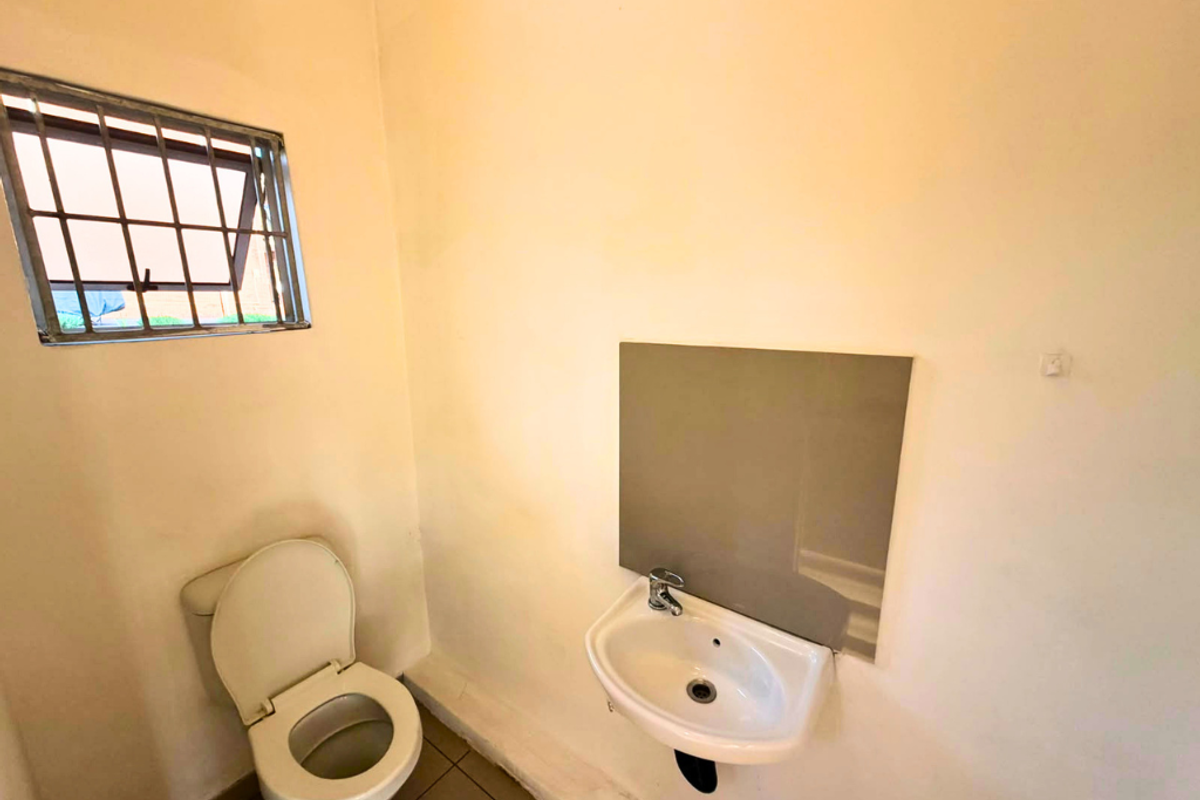 A compact, functional bathroom featuring a wall-mounted basin and toilet, finished with tiled flooring and a window with security bars.