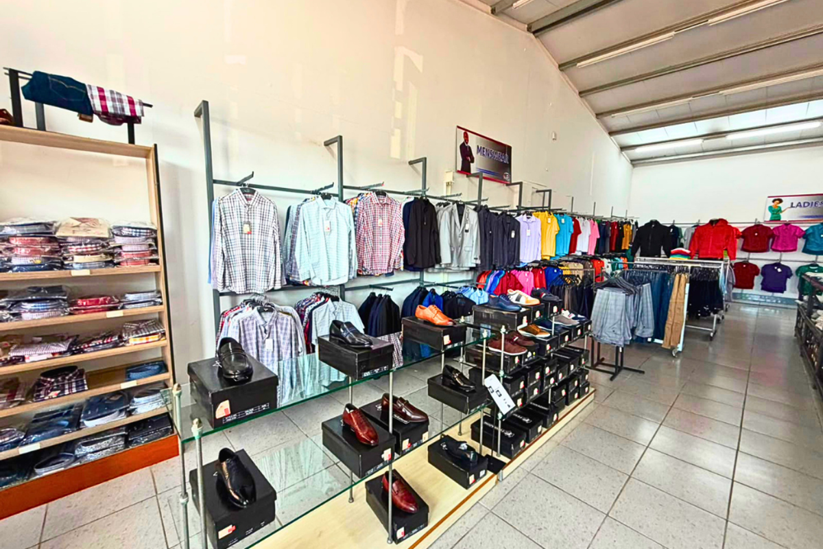 A commercial retail space featuring tiled flooring, exposed steel roof trusses, and extensive metal clothing display racks with integrated glass shelving.