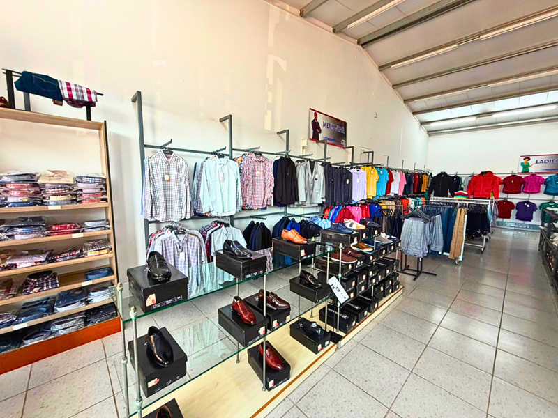 A commercial retail space featuring tiled flooring, exposed steel roof trusses, and extensive metal clothing display racks with integrated glass shelving.