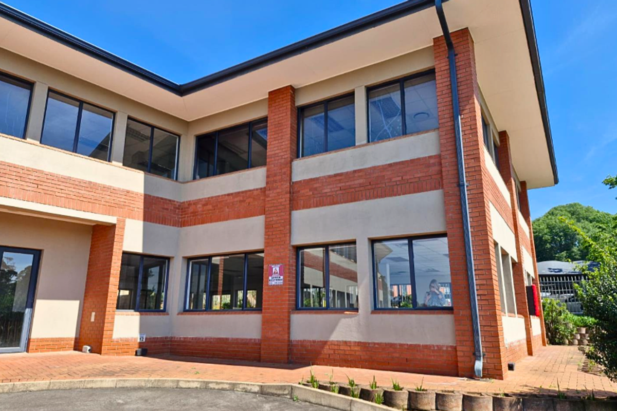 A modern two-story office building featuring a brick and plaster facade with large aluminium-framed windows and a paved entrance area.