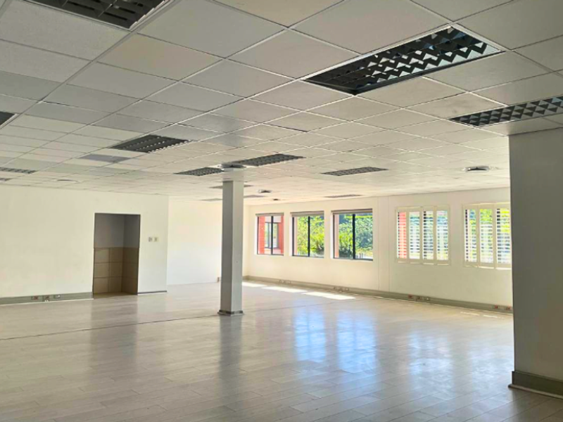 A spacious, open-plan office area featuring a suspended ceiling with recessed lighting, laminate flooring, and large windows with fitted shutters.