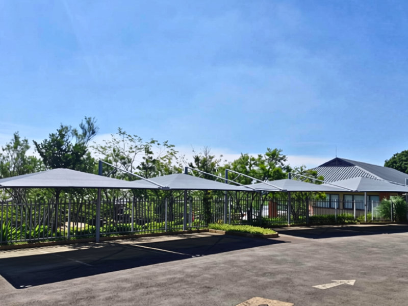 Outdoor parking area featuring a series of shade sail structures, asphalt paving, and secure metal perimeter fencing.