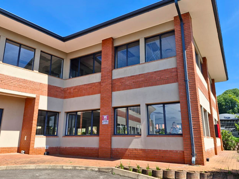 A modern two-story office building featuring a brick and plaster facade with large aluminium-framed windows and a paved entrance area.