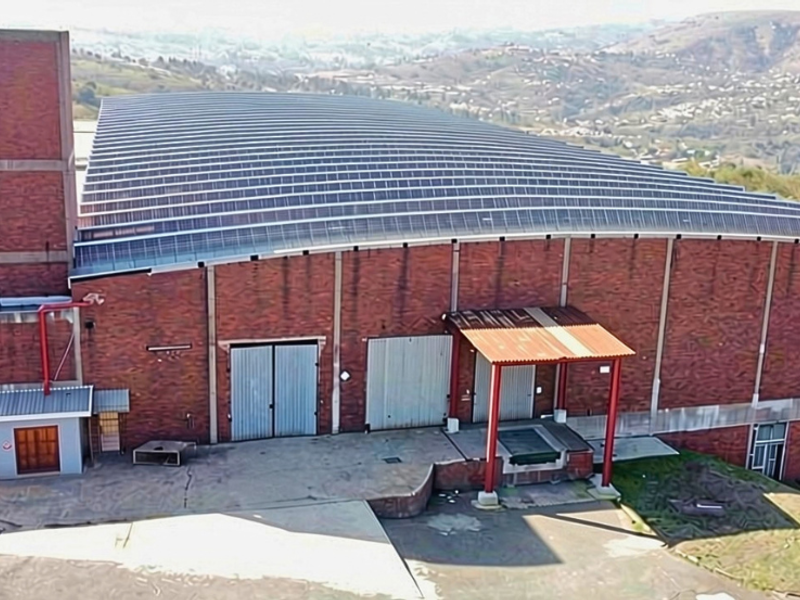 Aerial view of an industrial facility featuring a large brick warehouse with solar panels, loading bays, and a concrete yard area.