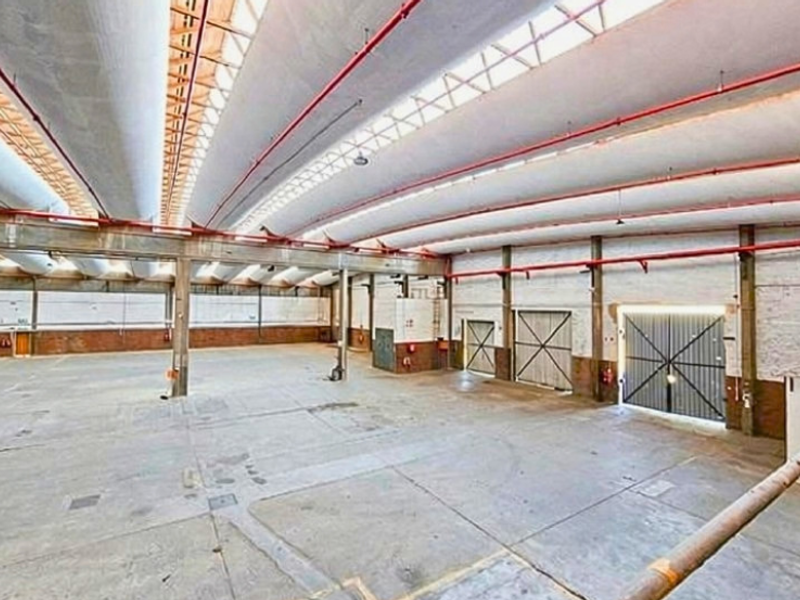 Spacious industrial warehouse featuring high ceilings with skylights, exposed fire sprinkler piping, concrete flooring, and large sliding access doors.